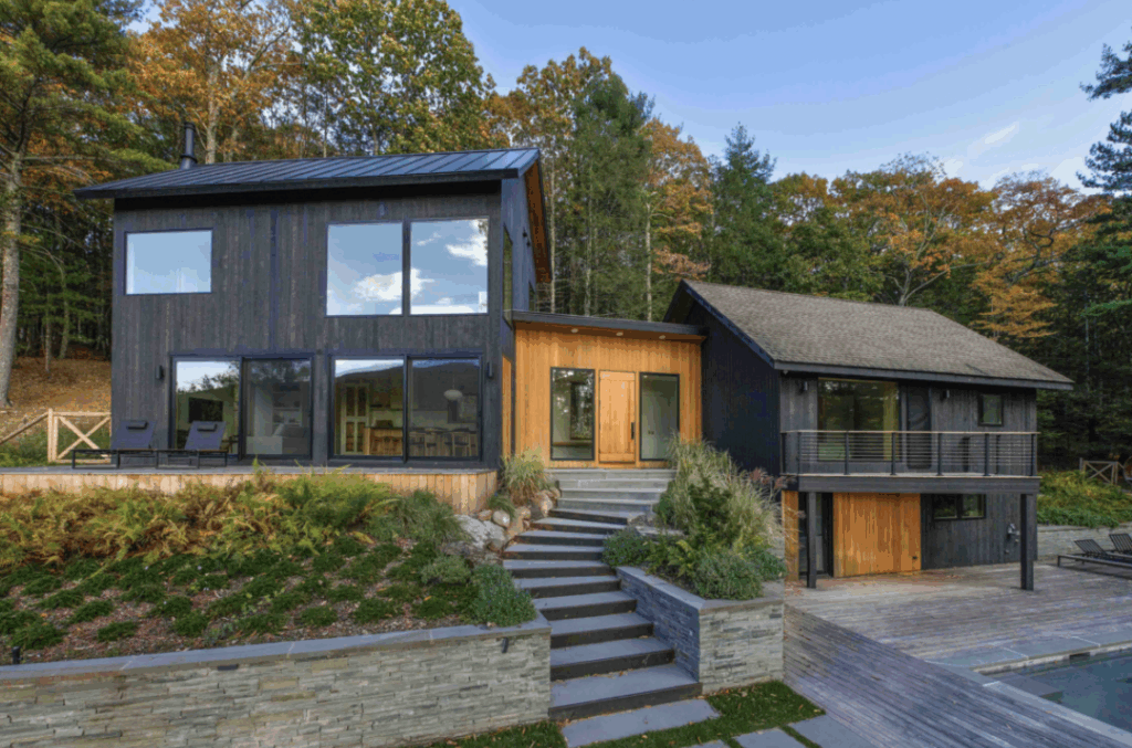 Modern two-story house with black wood siding and large windows, designed by custom home architects. Surrounded by autumn trees, stone steps and landscaping lead to glass double doors. A deck and pool are in the foreground.